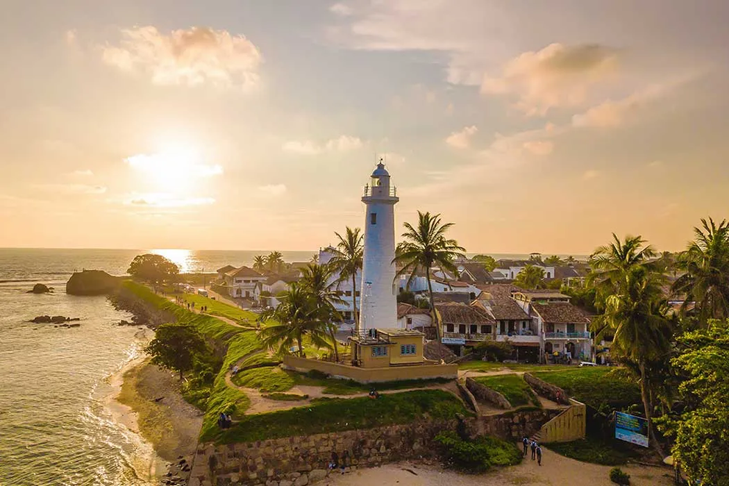 Galle lighthouse at sunset, with palm trees and the rooftops of the old fort along the coast