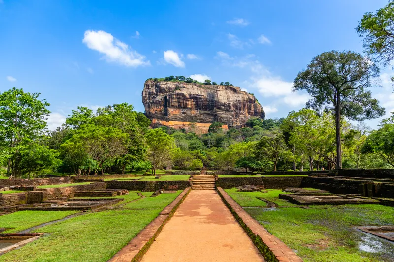 Sigiriya Rock Fortress rising above green gardens, with a red-earth path leading toward it under a bright blue sky